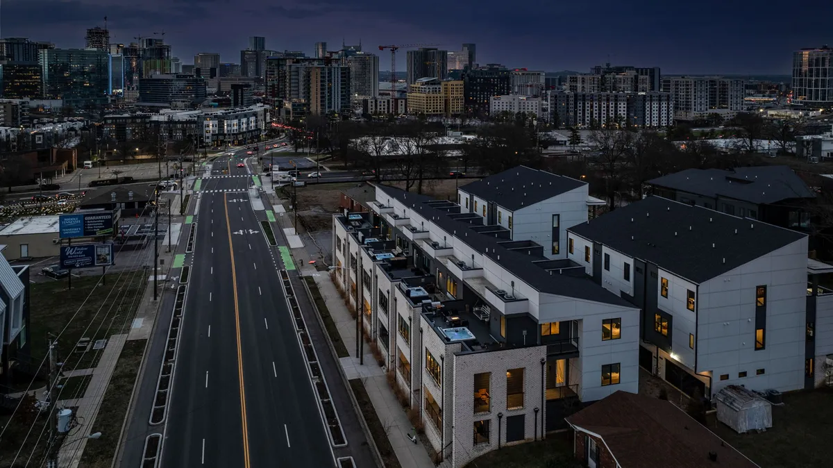 A row of modern townhouses illuminated at dusk, with the city skyline visible in the distance. The image captures the vibrant urban setting and hints at the exclusive rooftop patios each unit enjoys. The glow from the windows suggests a welcoming, lively community atmosphere.