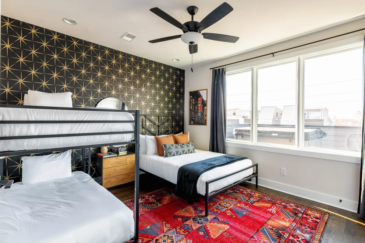 A cheerful, family-friendly bedroom featuring a queen bed and a bunk bed, with a colorful rug and playful star-patterned wallpaper. The window lets in natural light, and the design blends fun elements with contemporary style, making it suitable for both children and adults.