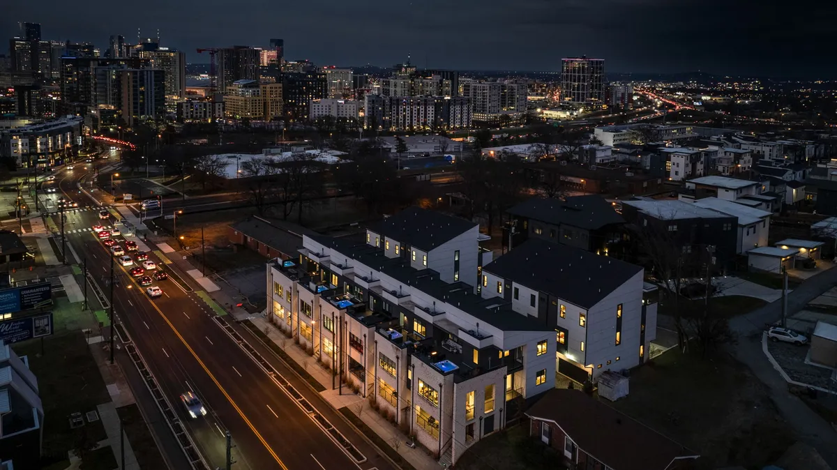 Modern townhouses line the street, with the Nashville city skyline rising in the background. Street lights and evening ambiance suggest urban vibrancy, while the rooftops hint at elevated spaces for relaxation and gathering.