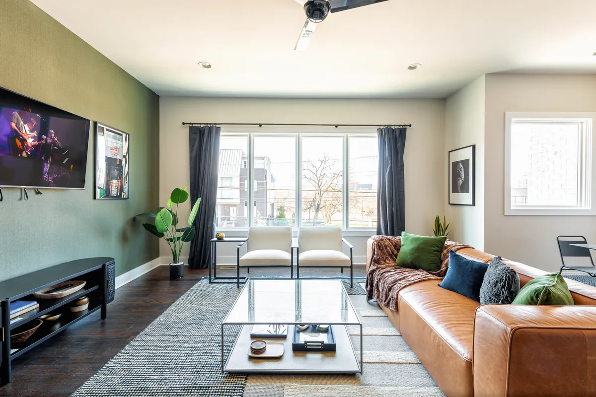 A sunlit living room featuring a leather sofa, stylish coffee table, large window allowing in natural light, and a TV. The design is both comfortable and sophisticated, suggesting a space ideal for gatherings or relaxing evenings.