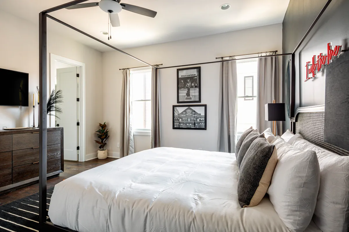 A luxurious king bedroom with a stylish four-poster bed, decorative cushions, wall art, and a TV. Sunlight streams into the room, creating a serene and comfortable atmosphere ideal for relaxation.